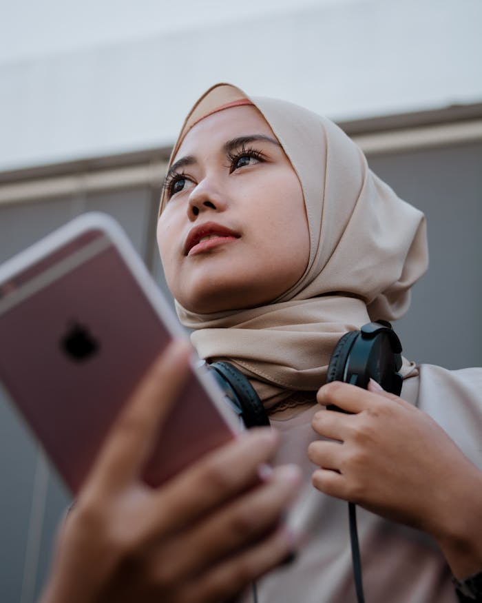 about-us Young woman in hijab using smartphone and headphones, embodying contemporary tech lifestyle.